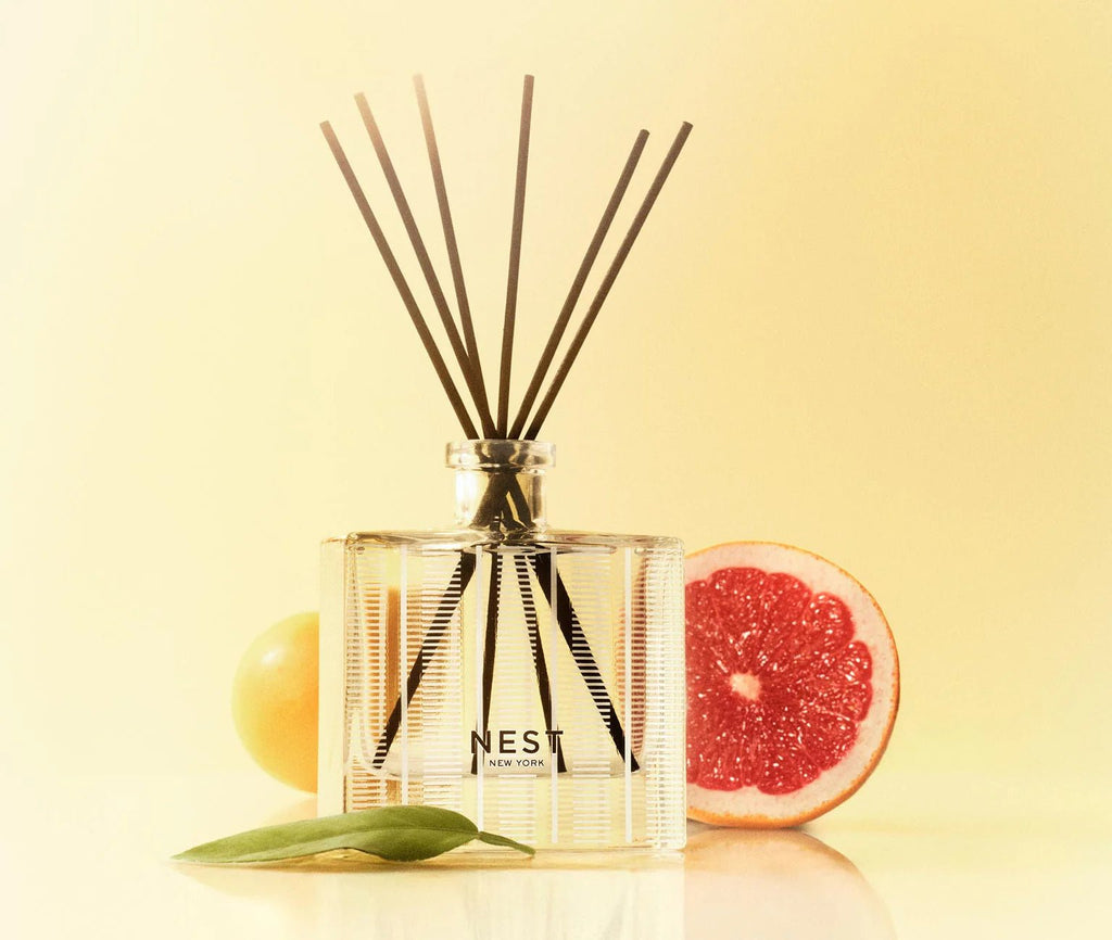 A Nest - Grapefruit Reed Diffuser by Nest is displayed next to a grapefruit half, a yellow sphere, and a green leaf, evoking fresh citrus notes against a soft yellow background.