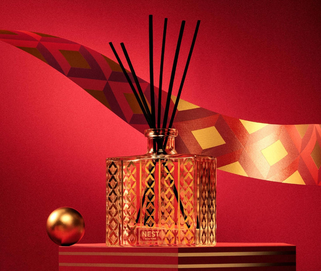 The Nest - Holiday Reed Diffuser, with its decorative glass bottle and black reed sticks, stands against a rich red background. It exudes a festive fruity aroma amidst a golden geometric ribbon and nearby gold sphere, creating an elegantly lit, luxurious holiday atmosphere.