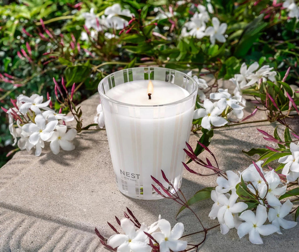 The Nest - Indian Jasmine Classic Candle by Nest rests on a stone surface, surrounded by blooming white jasmine flowers and green foliage, its glass holder highlighted in natural sunlight for a clean, fragrant burn.