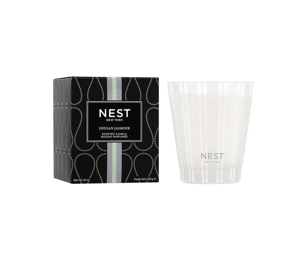 The Nest Indian Jasmine Classic Candle, in a white glass jar with vertical stripes and the Nest logo, sits beside its black, dotted box labeled "Indian Jasmine Scented Candle." It offers a clean burn and elegant presentation.