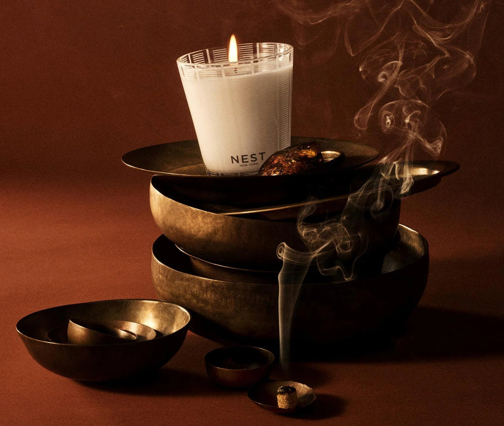 A white Nest - Moroccan Amber Classic Candle (Nest) burns atop stacked brass bowls, incense smoke wafting from a nearby small bowl, the scene set against a brown background.