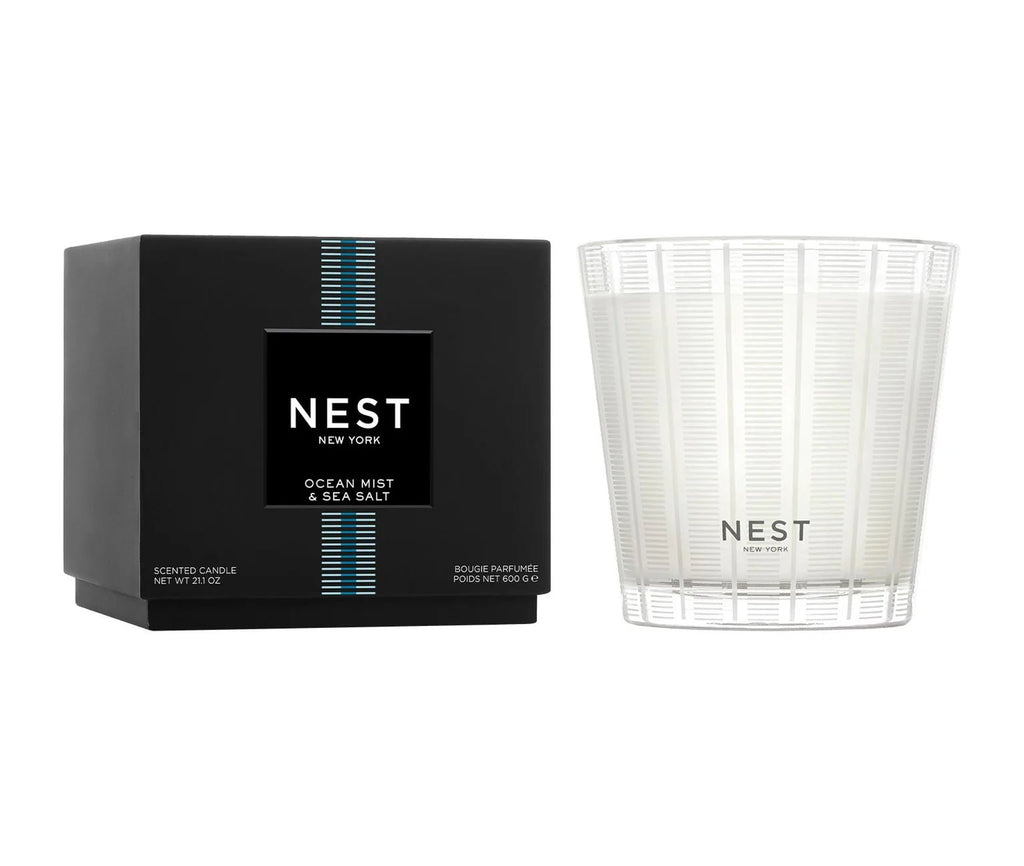 A black box with blue and white stripes labeled “Nest - Ocean Mist & Sea Salt 3-Wick Candle” sits next to a white patterned candle, both highlighting the fresh Ocean Mist & Sea Salt scent from Nest.