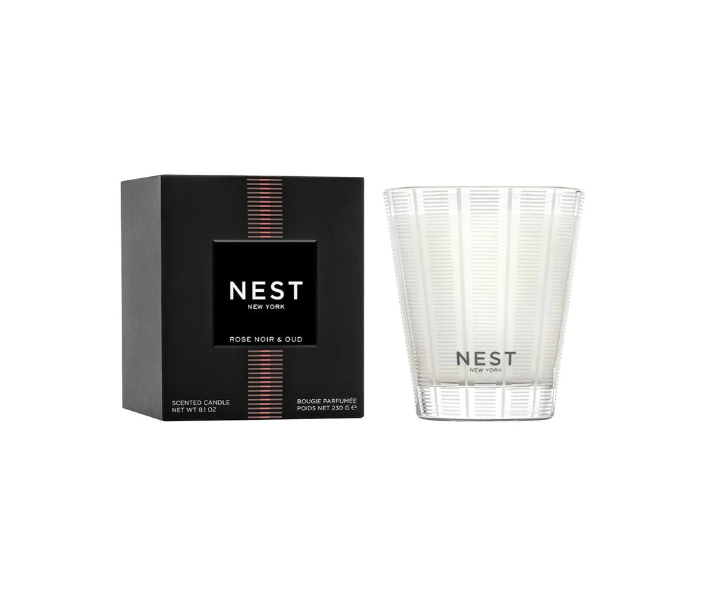 A Nest - Rose Noir & Oud Classic Candle from Nest is pictured in a clear, patterned glass next to its black box. The unused white candle features notes of Rose de Mai and smoky oud.