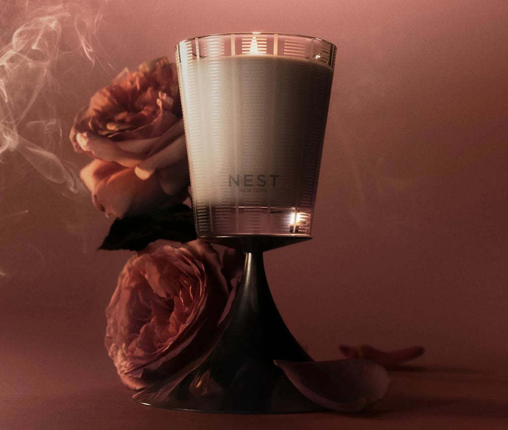 A lit Nest - Rose Noir & Oud Classic Candle in a glass holder sits on a reflective surface beside two pink Rose de Mai roses, with wisps of smoke and a warm, pink-toned backdrop evoking a cozy, elegant ambiance.