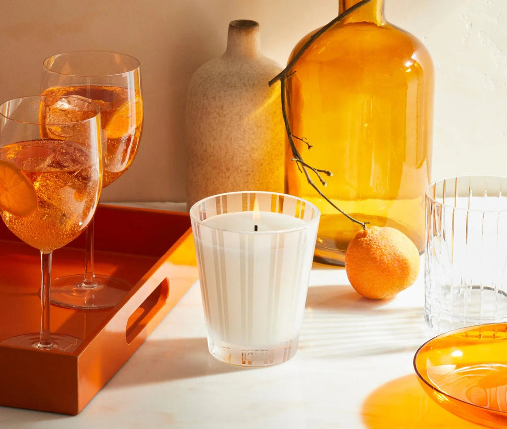 Two glasses with orange drinks, an orange tray, a Nest Sicilian Tangerine Classic Candle, a clear glass, an orange bottle with a branch, a beige vase, a Sicilian tangerine, and an orange dish are arranged on a white surface in warm sunlight.
