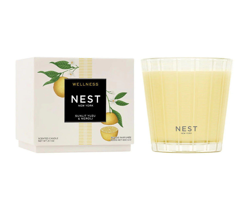 The Nest Sunlit Yuzu & Neroli 3-Wick Candle sits in a ribbed glass holder beside its white box, which features colorful citrus fruit artwork and Nest branding.