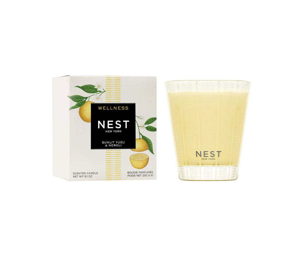 A Nest - Sunlit Yuzu & Neroli Classic Candle in a clear glass jar sits next to its box, which features vibrant yuzu fruit and leaf illustrations, reflecting the fresh citrus scent of this signature Nest candle.