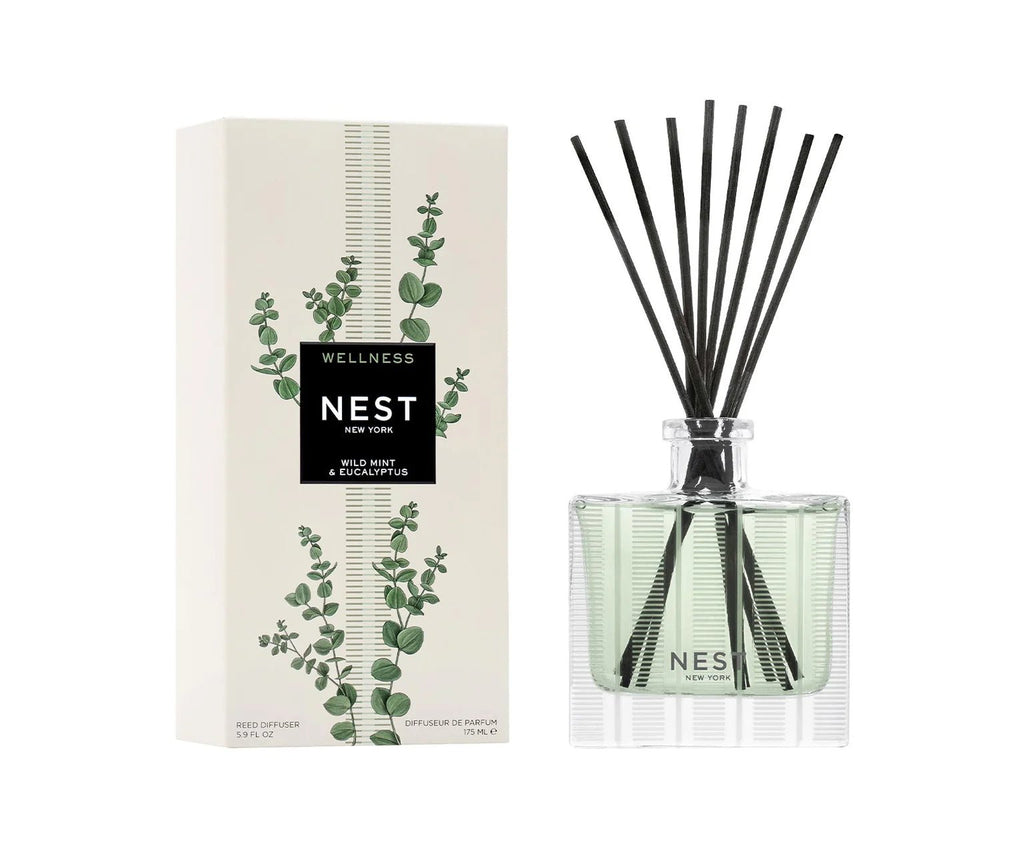 A Nest - Wild Mint & Eucalyptus Reed Diffuser from Nest in a clear glass bottle with black reeds sits beside its white box, which features green eucalyptus designs and is labeled “Wellness.”.