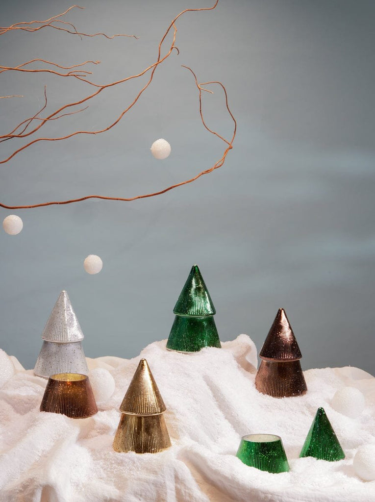 The Paddywax 7oz Cypress + Fir Mercury Trees in green by Paddywax sit on a snowy white surface with bare branches and white hanging balls, complemented by mercury glass candles against a soft blue background for a festive look.