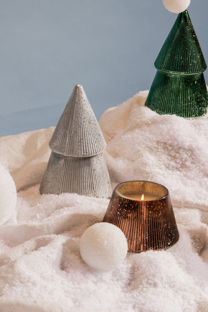 The Paddywax 7oz Cypress + Fir Mercury Trees in silver and green stand on white artificial snow, while a lit ribbed brown glass candle holder complements large white snowballs, enhancing the metallic display.