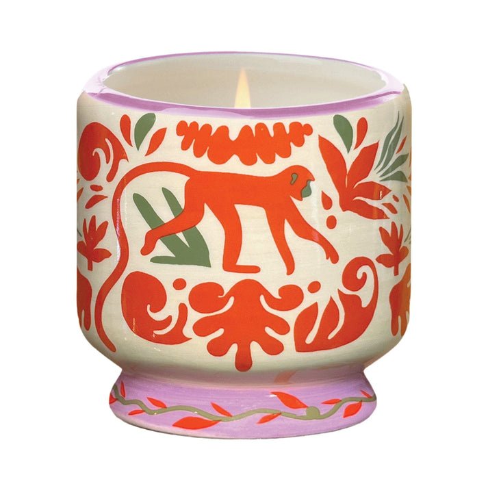 The Paddywax A Dopo 8 oz Handpainted Coconut + Amber Ceramic candle features a cream-colored holder with red and green hand-painted patterns, including a red monkey and leafy designs, showcased against a plain white background.