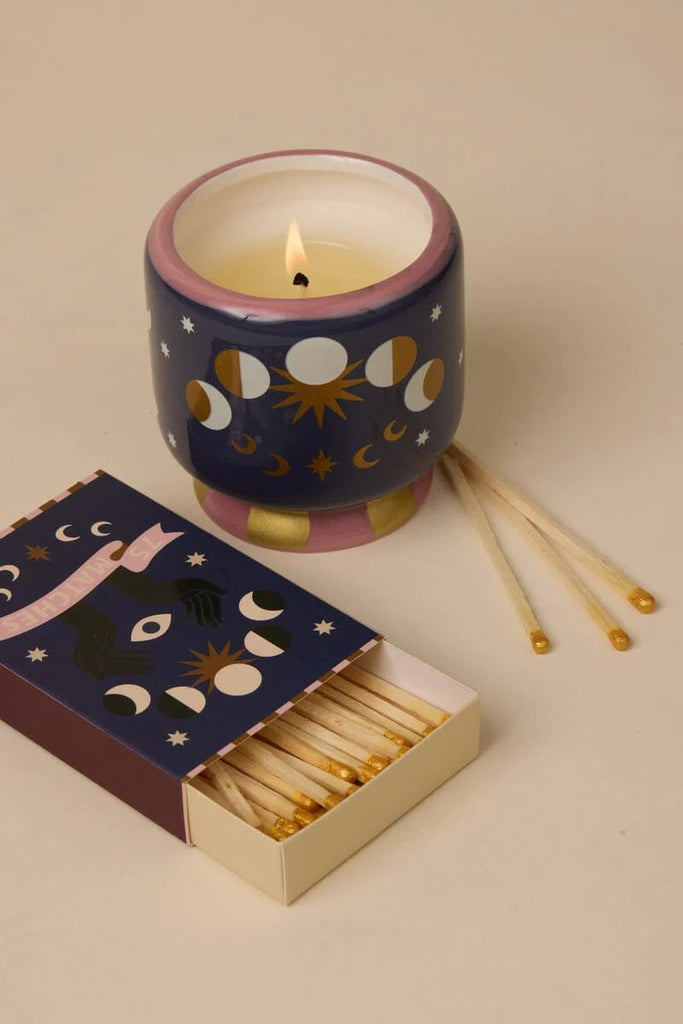 A Paddywax A Dopo 8 oz handpainted Moon Phases ceramic candle with a Jasmine & Rose scent sits lit on a beige surface. Beside it is an open box of illustrated matches with celestial motifs, and a few matches are scattered around.