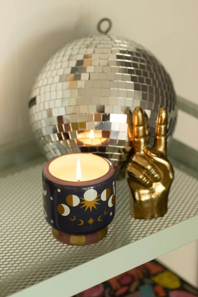 The Paddywax A Dopo Jasmine & Rose candle, in an 8 oz hand-painted Moon Phases ceramic, sits next to a disco ball and gold peace sign on a metal shelf, filling the air with its scent and creating a cozy ambiance.