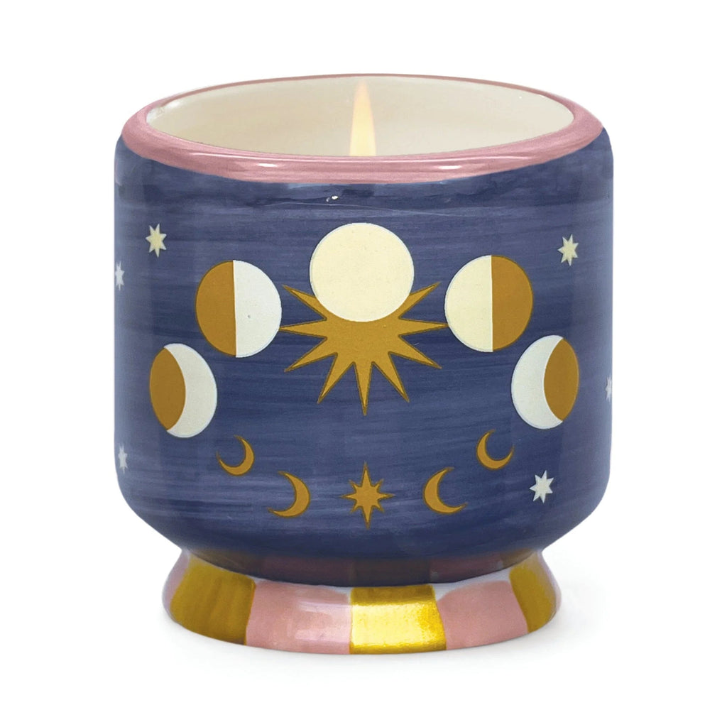 The Paddywax Moon Phases decorative ceramic candle features celestial designs on a dark blue background with a pink rim and base striped in pink and gold. It holds hand-poured soy wax, releasing the Jasmine & Rose scent.