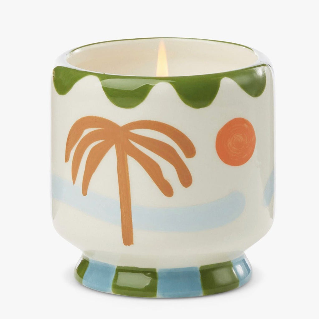 The Paddywax - A Dopo Lush Palms Handpainted 8oz Candle features a soy wax candle in a hand-painted ceramic vessel with a brown palm tree, orange sun, green waves, and blue stripes on an off-white background. The base displays blue and green checkered patterns.