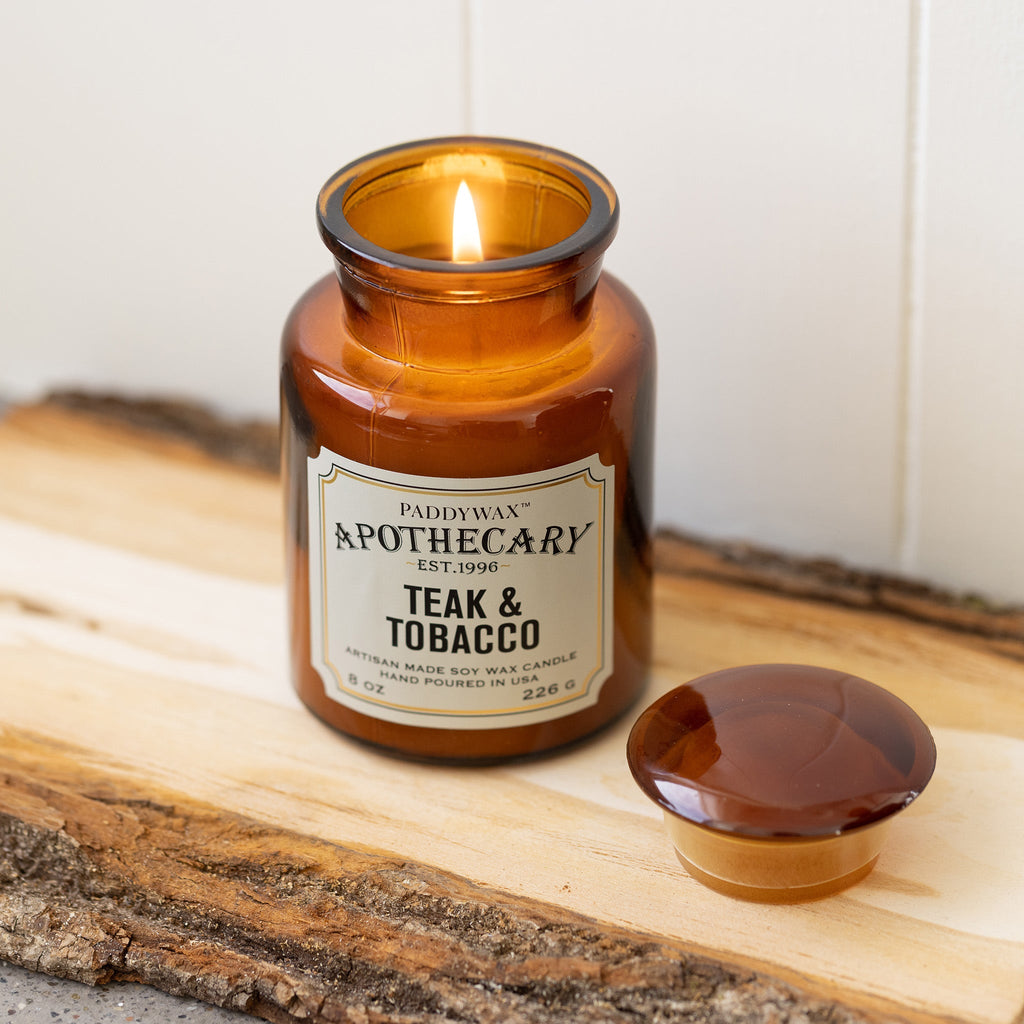 A lit 8oz Paddywax - Apothecary - Teak & Tobacco candle in an amber glass vessel sits on a wooden surface. The lid is beside it, emitting a warm glow and releasing a luxurious fragrance.