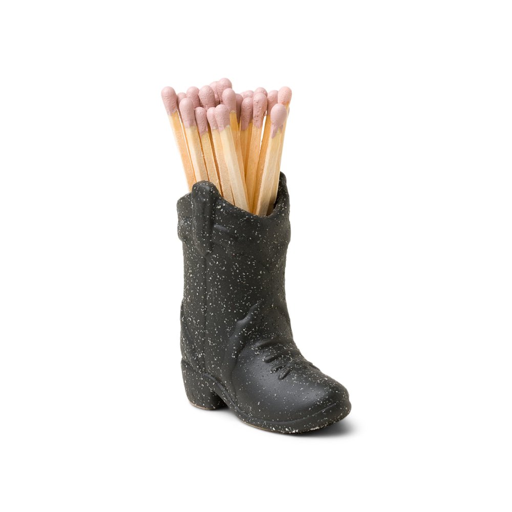 The Paddywax Black Cowboy Boot Match Holder is a small ceramic container with a textured, speckled finish, perfect as a Nashville gift, and it holds several wooden matches with pink tips.