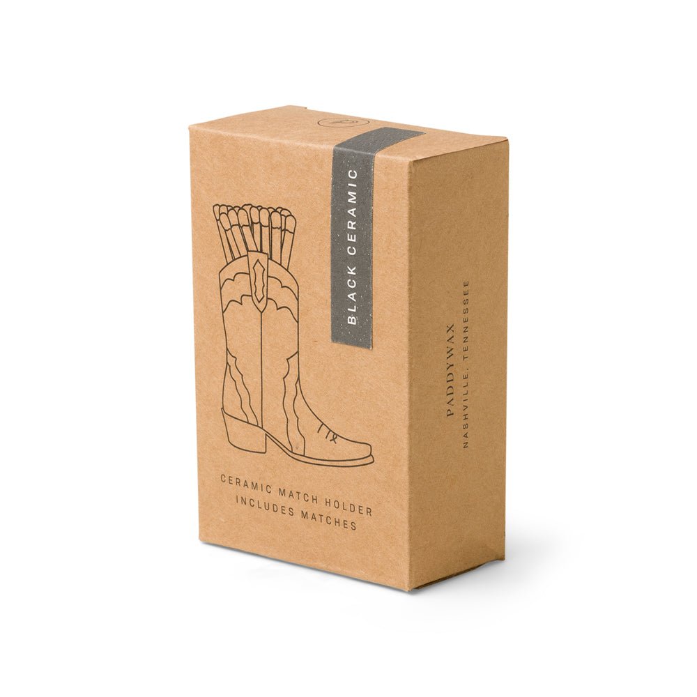 The Paddywax Black Cowboy Boot Match Holder is a charming Nashville gift. It features a black line illustration of a ceramic cowboy boot filled with matches on its brown box, labeled Black Ceramic and Ceramic Match Holder Includes Matches on the side.