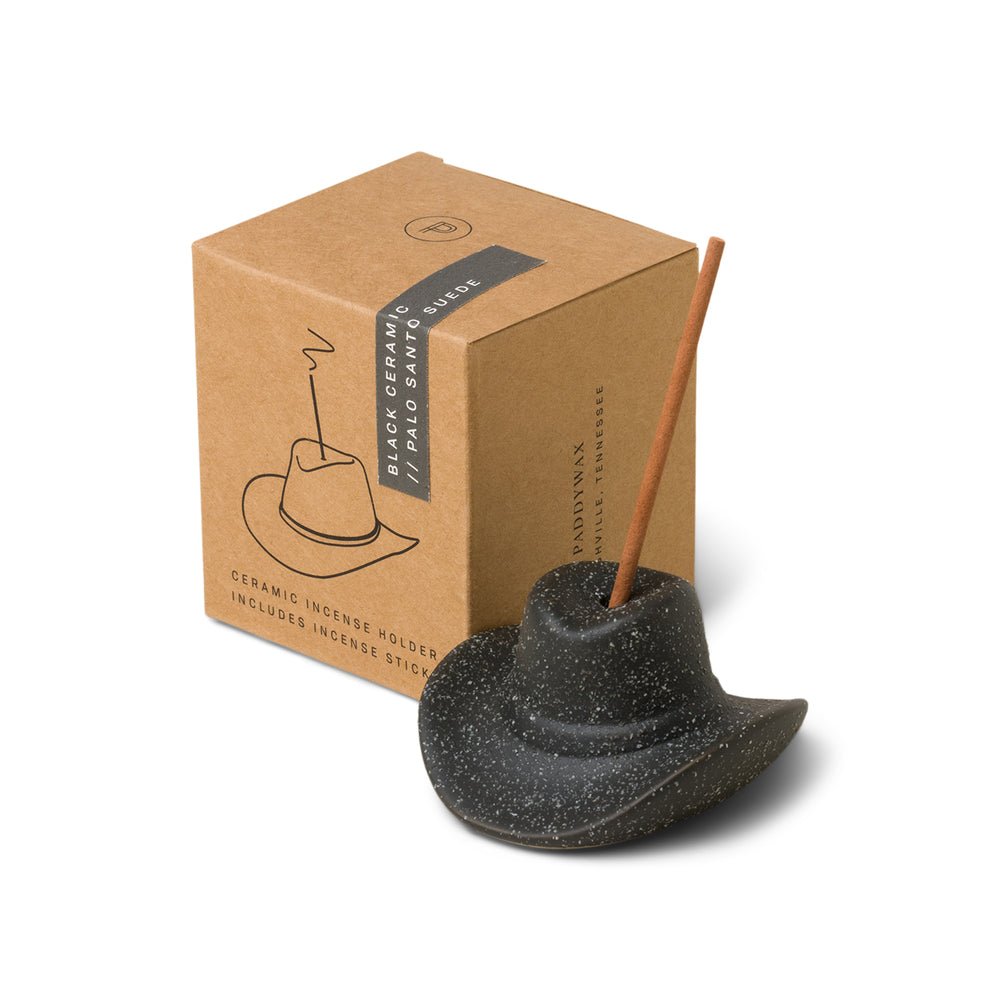 The Paddywax Black Cowboy Hat Ceramic Incense Holder offers a Western flair, featuring a lit stick in its black brim. It comes with brown packaging illustrated with the product and Black Ceramic Incense Holder text—ideal for Palo Santo Suede enthusiasts.