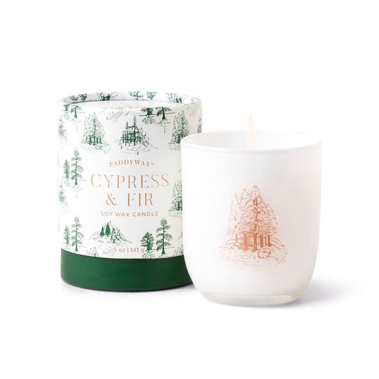 The Paddywax Cypress & Fir 5oz white opaque glass candle is elegantly showcased next to its decorative cylindrical packaging. The frosted fir needle design alongside green tree illustrations adds a subtle pine tree charm to both the candle and its packaging.