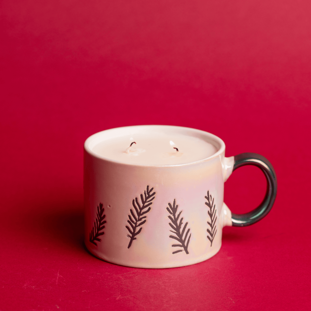 The Paddywax Cypress & Fir White Ceramic Mug Candle features a fern-adorned design against a red background. Its unused white candle, with two wicks and an elegant dark handle, makes it a perfect dual-purpose gift.