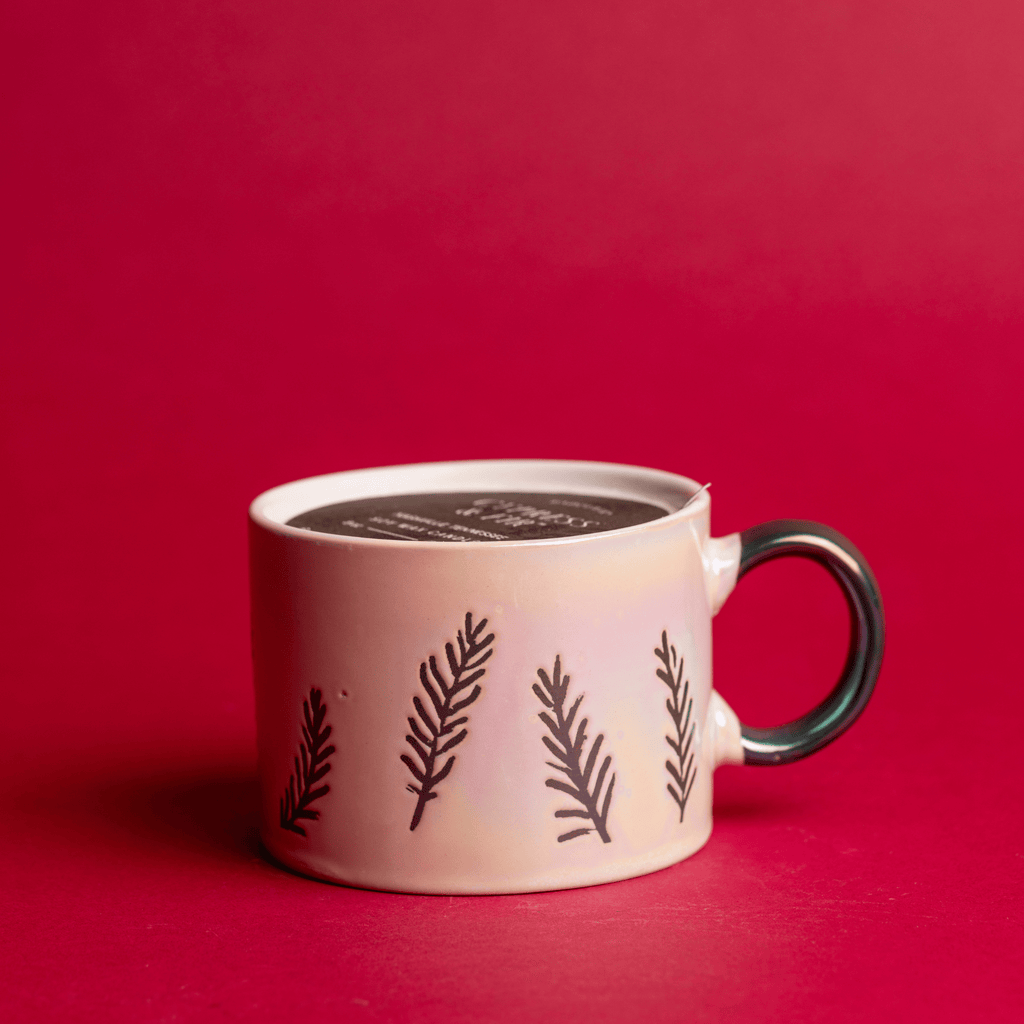 A white ceramic mug containing a Paddywax Cypress & Fir candle sits on a red background, featuring black leaf-like patterns. Its elegant handle complements the design, making it an ideal dual-purpose gift for any occasion with a smooth surface hinting at coffee or hot chocolate.