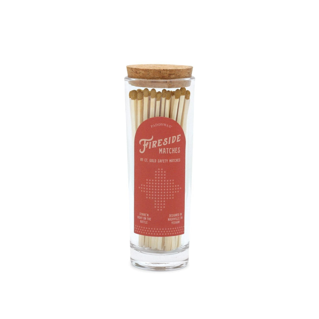 The Paddywax - Fireside Safety Matches - Gold come in a glass jar with a cork lid, containing long wooden matches with gold tips. The jar features a red label with white text and a minimalist campfire design, making it an ideal candle companion with vintage charm.