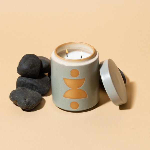 The Paddywax Form Ocean Rose + Bay 12oz Candle, in a ceramic vessel with a geometric design, sits on a beige surface, its fragrance gently filling the air. The lid is set aside, with smooth black stones artfully arranged next to this exquisite planter candle.