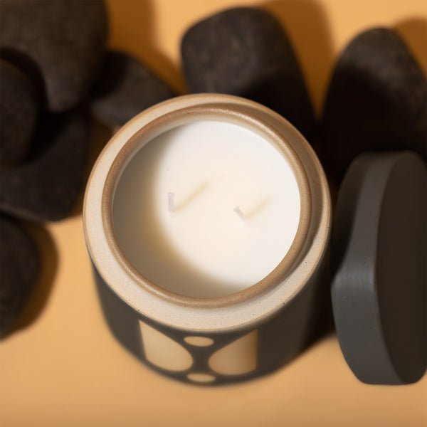 Top view of a lit Paddywax - Form - Palo Santo Suede 12oz Candle in a decorative ceramic vessel, surrounded by smooth black stones on a beige background. The two-wick candle has an open lid beside it, releasing a soothing fragrance.