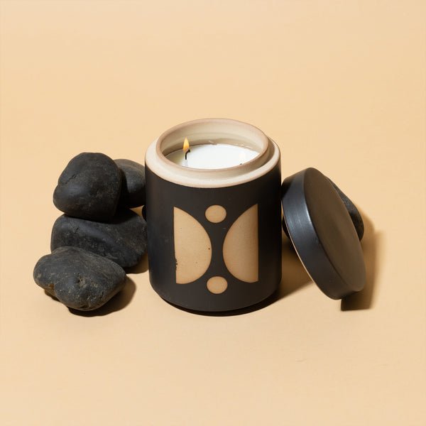 A small, gentle flame from the Paddywax - Form - Palo Santo Suede 12oz Candle in a black jar with geometric designs releases a delicate scent beside smooth black stones on a beige background. The ceramic vessels lid is placed slightly aside.