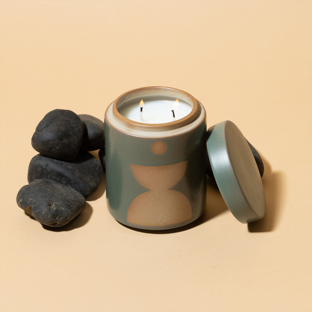 The Paddywax - Form - Spanish Moss 12oz Candle features a ceramic vessel with geometric patterns and two wicks, sitting on a beige surface. The jar lid is placed aside, surrounded by dark stones, adding elegance and fragrance to any setting.