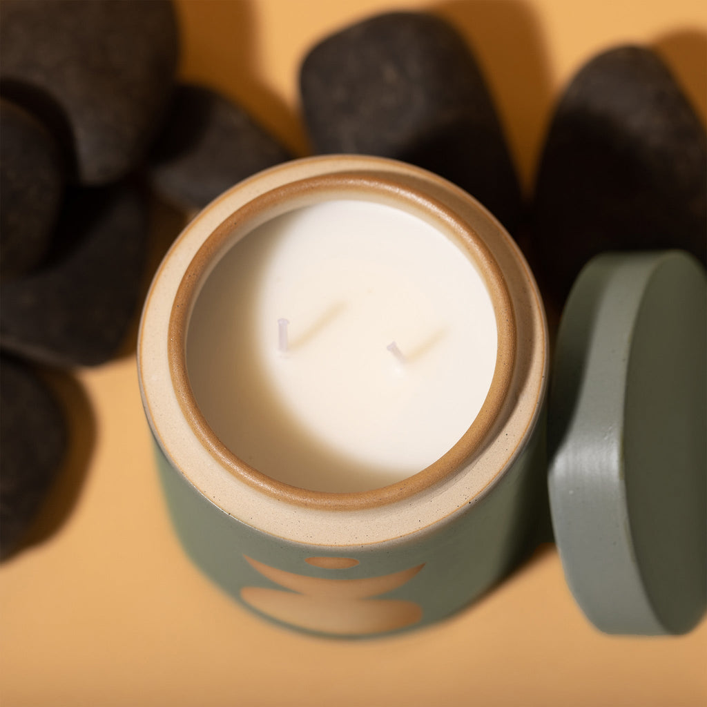 Overhead shot of the Paddywax - Form - Spanish Moss 12oz Candle, a green ceramic vessel with two wicks, surrounded by black stones on a light brown surface. The lid is ajar beside the jar, emitting a subtle fragrance.