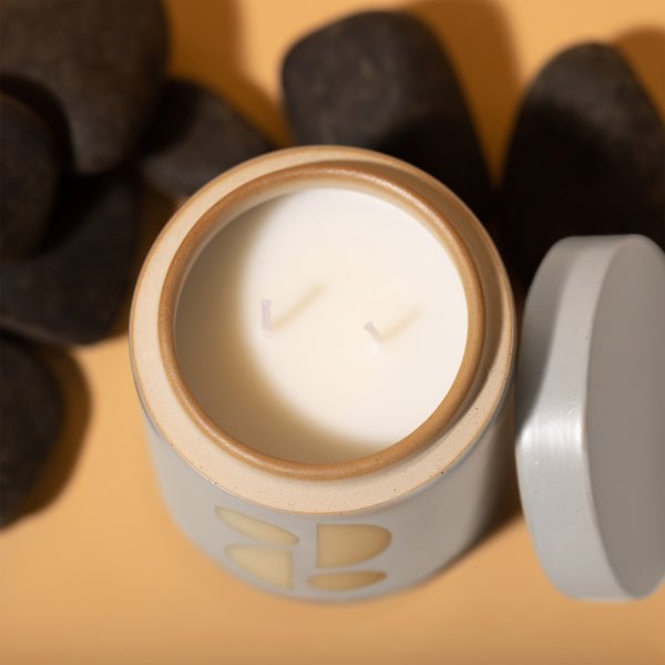 A white 12oz Tobacco Flower candle by Paddywax, featuring two wicks in a geometric ceramic vessel, sits on a beige surface. The jar lid hints at its garden fragrance, while black stones lie scattered in the background.