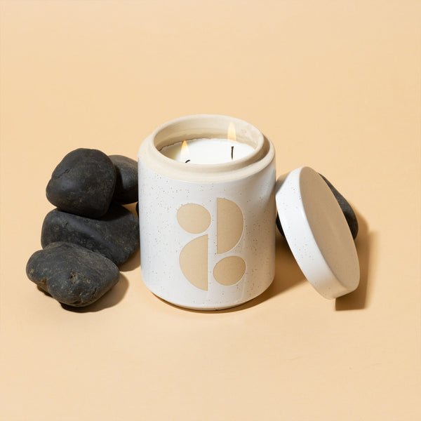 The Paddywax - Form Tobacco Flower 12oz Candle, a white ceramic jar with a minimalist design and multiple wicks, sits lidless against a beige background. Surrounded by smooth black stones, it exudes a garden-inspired fragrance.