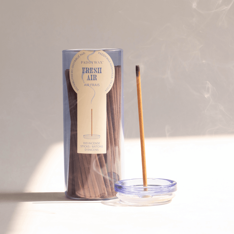 A Paddywax Haze Fresh Air Travel Incense cylindrical container sits beside a burning stick in a sleek glass vessel, with delicate smoke curling upward and filling the air with soothing fragrance against a soft, neutral backdrop.