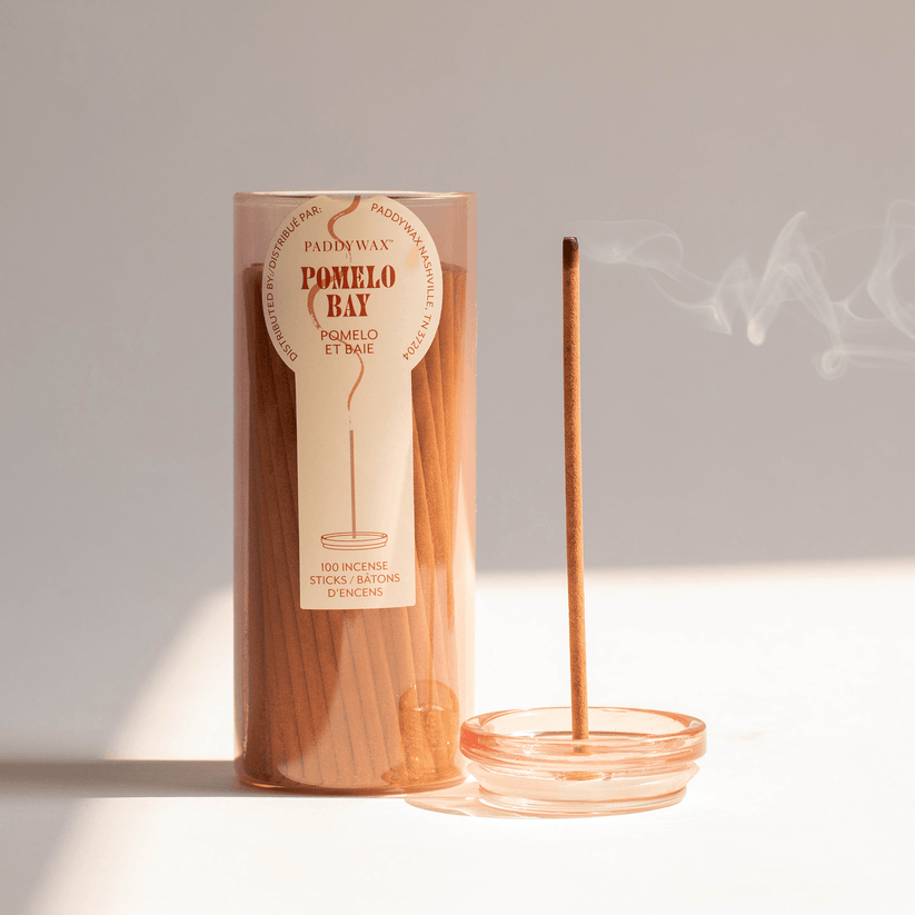 A cylindrical Paddywax - Haze - Pomelo Bay Travel Incense container, from the brand Paddywax, holds an incense collection. An incense stick stands in a glass holder, releasing smoke against a neutral backdrop, highlighting popular fragrances.