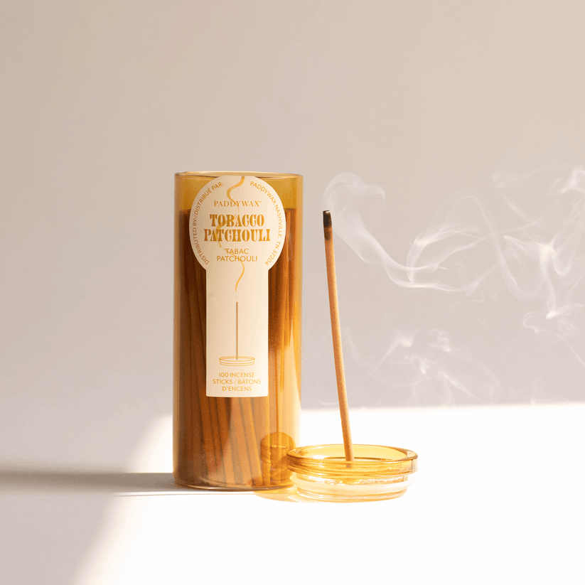 A cylindrical amber container labeled Paddywax - Haze - Tobacco Patchouli Travel Incense features a candle logo. A used match lies on the glass cap beside it, wisps of smoke curling up like incense. Natural light from the right casts soft shadows.