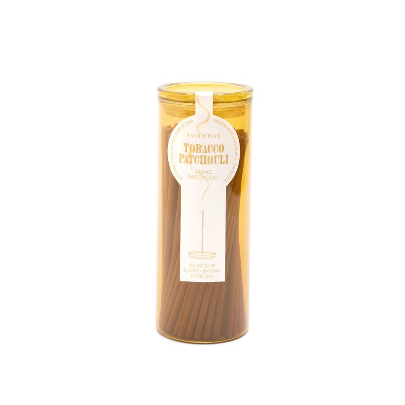 The Paddywax Haze Tobacco Patchouli Travel Incense comes in a cylindrical vessel with a transparent lid, showcasing brown incense sticks. A white tag with text and logo adds charm to the elegant packaging, promising an enchanting fragrance experience.