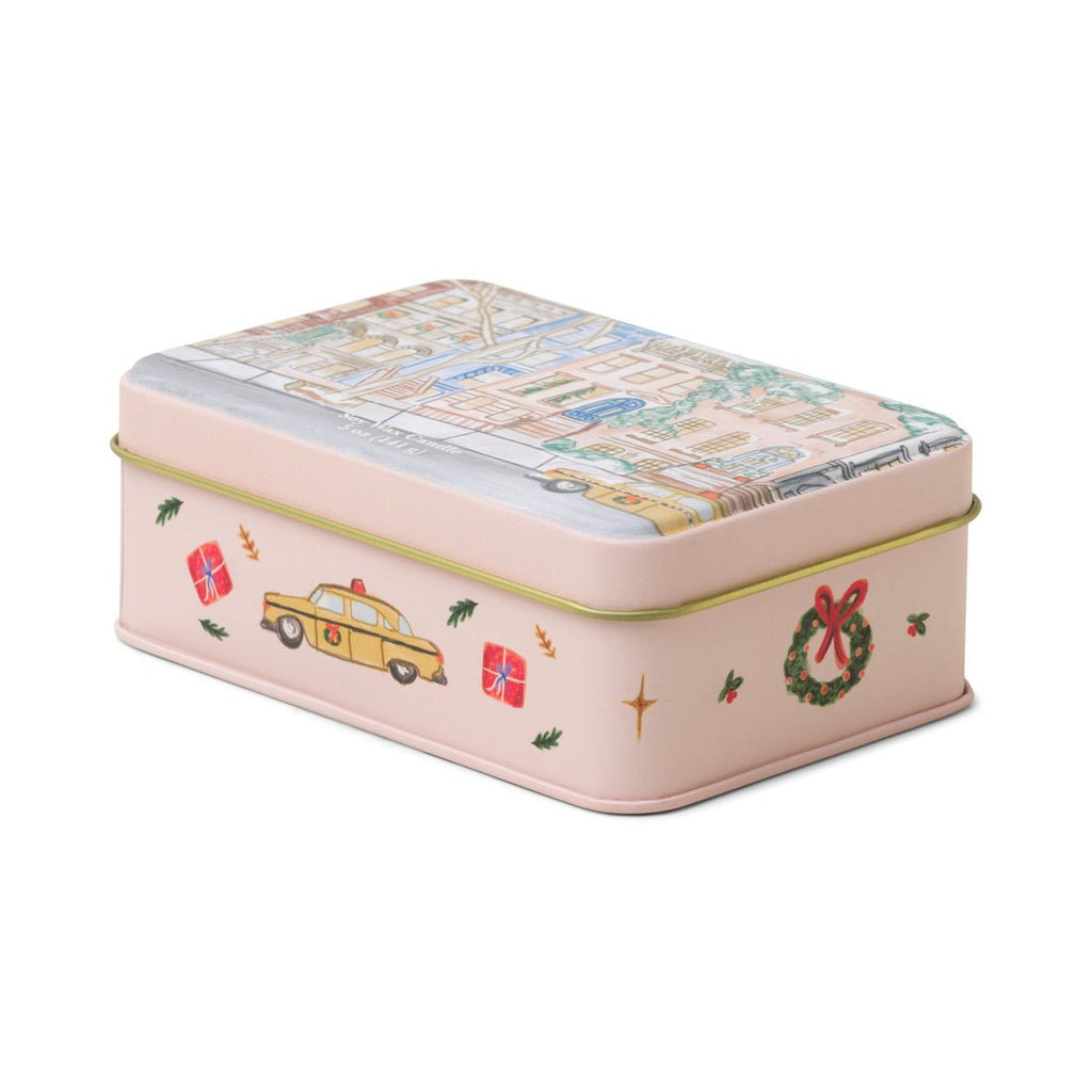 The Paddywax Holiday Candle Tin features a map-illustrated lid. The rectangular tin box showcases festive designs, including a yellow car, green wreath with a red bow, and red presents on a light pink background. Its perfect for gifting holiday candles with tangerine and clove fragrances.