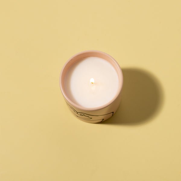 A Paddywax Impressions Love Ya candle in a pink ceramic holder rests on a light yellow surface, emitting a tobacco vanilla scent. Its single flame flickers softly, casting shadows to the right. The colorful matte finish provides an elegant touch to the serene setting.