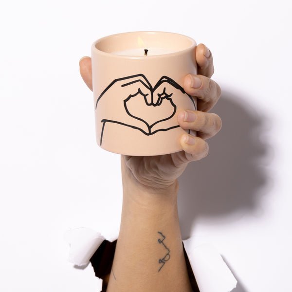 A hand holds a lit Paddywax Impressions candle in Tobacco + Vanilla with Love Ya inscription, featuring two hands forming a heart design. A tattooed wrist emerges from a tear, and the candle emits cozy notes of cinnamon nutmeg.