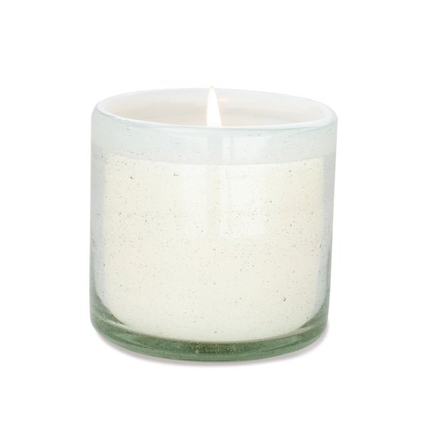 A lit Paddywax La Playa Amber + Coconut 9oz candle in a hand-blown glass container against a white background, softly glowing and casting a warm light that evokes beach vibes.