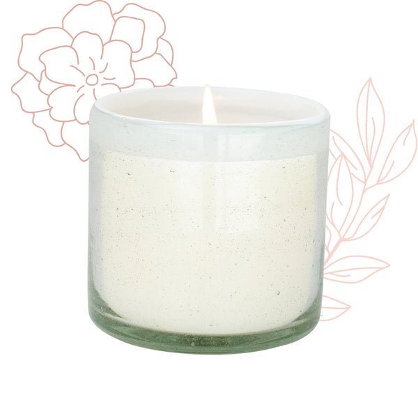 The Paddywax La Playa Amber + Coconut 9oz Candle is elegantly presented in a hand-blown recycled glass container, with a lit white candle. The background showcases soft peach outline drawings of flowers and leaves, enhancing the subtle beach vibes.