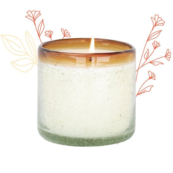 The Paddywax La Playa Orange Blossom 9oz candle features a lit candle in a textured glass holder crafted from hand-blown bubble glass with a brown rim, surrounded by minimalist orange floral illustrations on white, highlighting the skill of Mexican artisans.