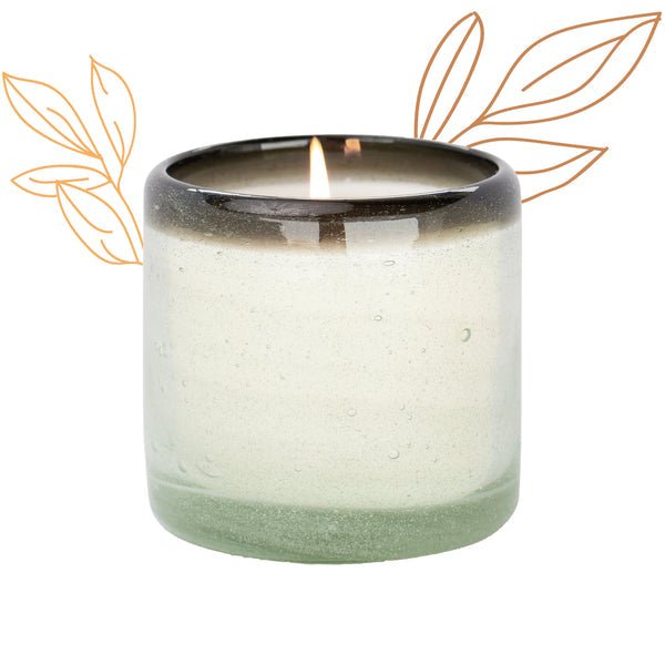 The Paddywax La Playa Vanilla Rosa 9oz Candle sits in a hand-blown recycled glass holder with a gradient from light to dark. Its white background is enhanced by simple orange leaf line drawings on each side, emphasizing the elegance of the vessel.