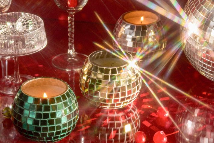 The Paddywax Magique 10 oz Green Disco Ball candle in Tapac & Pine sits on a shiny red surface, surrounded by mirror ball candles, glassware, and red berries for a festive atmosphere with sparkling reflections and light flares.