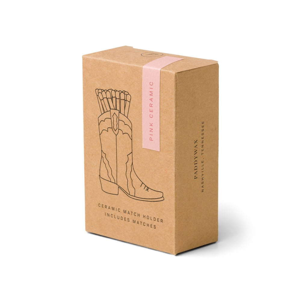 A brown box labeled Paddywax - Pink Cowboy Boot Match Holder contains a vintage-inspired ceramic match holder with matches. The cowboy boot illustration hints at the match holders Western design.