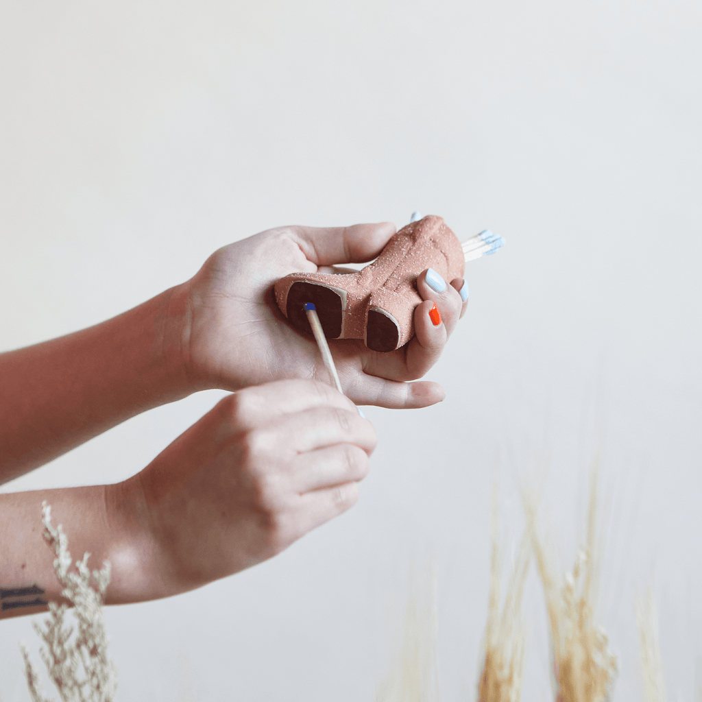 A person with orange and blue nails holds the Paddywax Pink Cowboy Boot Match Holder, inserting a thin stick to capture a Western vibe, while dried grasses softly blur in the background.