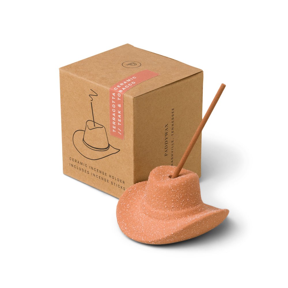 A ceramic incense holder shaped like a cowboy hat sits with an incense stick inserted, beside a brown box labeled Teakwood Tobacco & Leather, Paddywax, which features a line drawing of the Paddywax - Terracotta Cowboy Hat Ceramic Incense Holder.