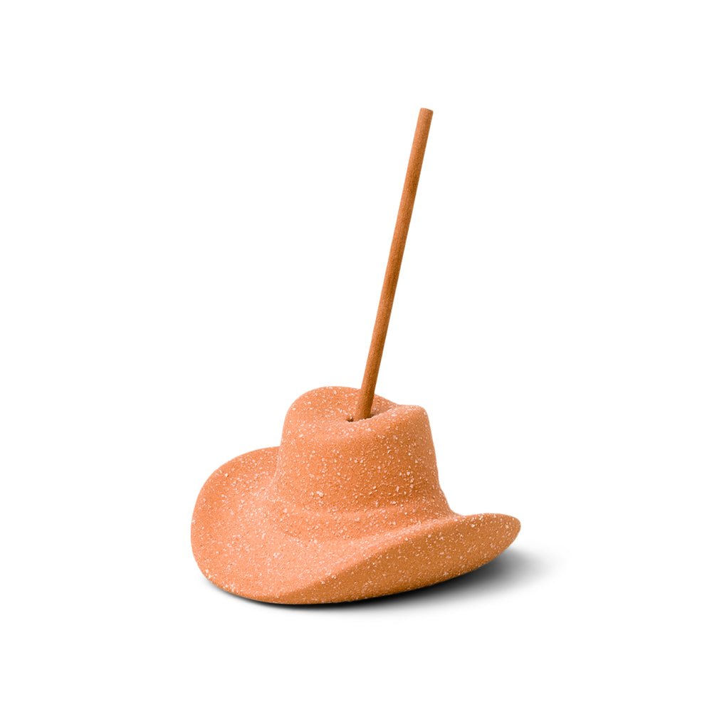 A cinnamon stick stands upright in a textured, light brown base, the Paddywax - Terracotta Cowboy Hat Ceramic Incense Holder, against a white background evoking the rich scent of Teakwood Tobacco and tamarind candy.